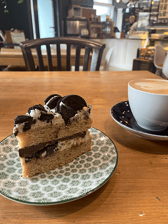 Kaffee & Kuchen im Matildas – Imbolc-Workbook fertig! Ein Teller mit Oreo-Cookie-Kuchen und ein Cappuccino im Matildas – Feiermoment nach der Fertigstellung des Imbolc-Workbooks