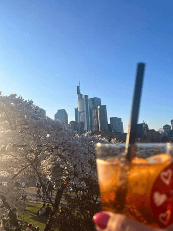 Aperol Spritz vor Frankfurter Skyline und Kirschblüten Ein Glas Aperol Spritz wird vor der Frankfurter Skyline und einem blühenden Kirschbaum in die Sonne gehalten