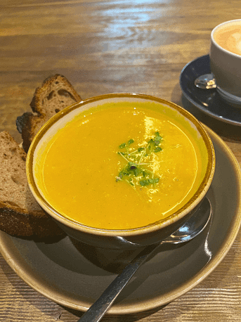 Kürbissuppe, Brot und Cappuccino in Matildas Kitchen Ein Teller Kürbissuppe mit Brot und ein Cappuccino auf einem Holztisch – warmer Genussmoment