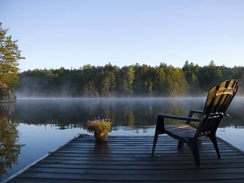 Ein leerer Stuhl auf einem Steg am See bei Sonnenaufgang – ein Symbol für Einkehr, Stille und Verbindung mit dem inneren Raum, wie in einer AKASHA-Lesung.