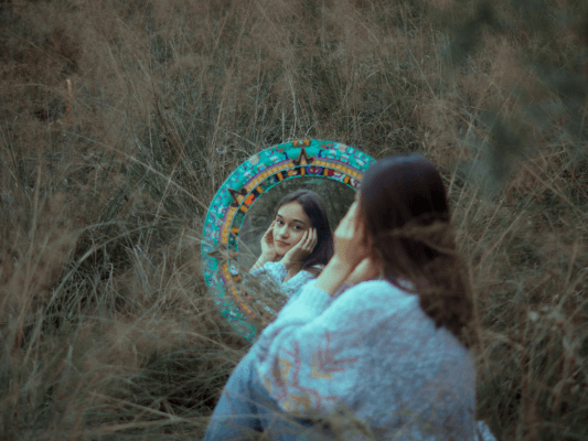 Junge Frau sitzt auf einer Wiese und betrachtet sich im Spiegel – Sinnbild für Selbstannahme Junge Frau sitzt auf einer Wiese und betrachtet sich im Spiegel - Sinnbild für Selbstreflexion und Selbstannahme