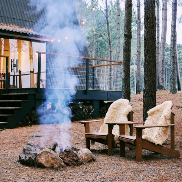 Zwei Holzstühle mit Fellauflagen vor Lagerfeuer mit moderner Hütte im Hintergrund