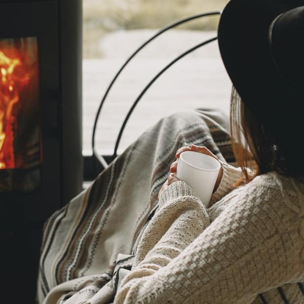 Frau mit Hut und Tasse in der Hand sitzend vor Kamin
