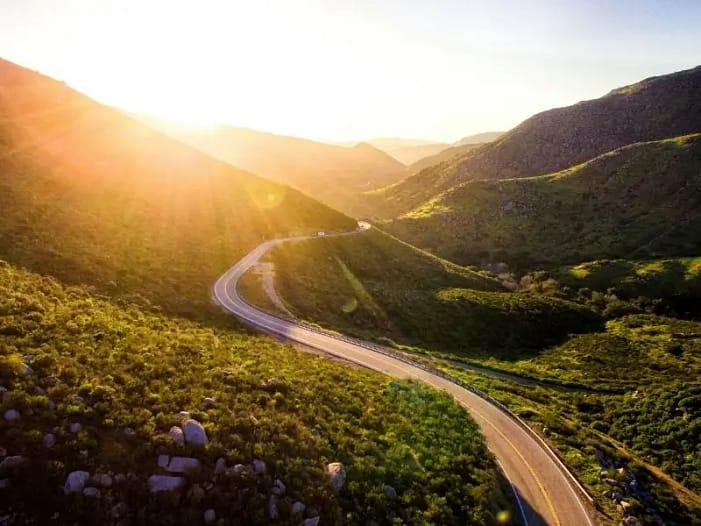 Sich windende Straße im Sonnenlicht - der individuelle Weg zur Lebensaufgabe Luftaufnahme einer sich windenden Straße durch grüne Hügel im goldenen Sonnenaufgangslicht - Symbol für den individuellen, nicht geradlinigen Weg zur eigenen Lebensaufgabe