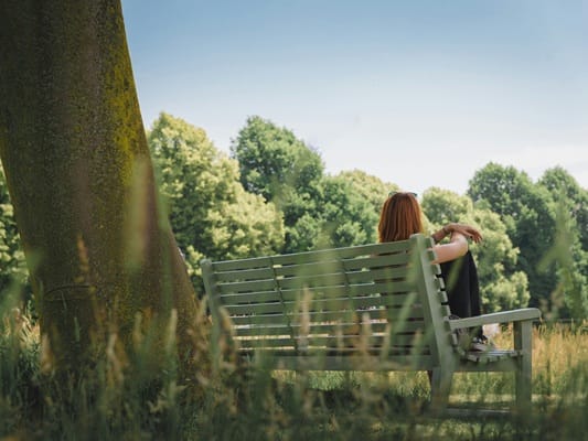 Frau sitzt auf einer grünen Bank unter einem Baum, blickt in die Natur – Symbol für Selbstreflexion & innere Ruhe