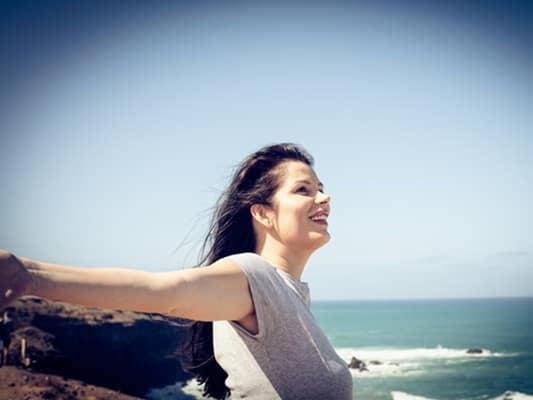 Frau am Meer mit Wind in den Haaren, blickt lächelnd in die Ferne – Symbol für Loslassen, Freiheit und emotionale Balance