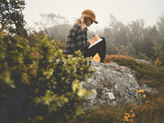 ine Frau sitzt ruhig auf einem großen Stein im Grünen und schreibt – ein Moment der Reflexion, um das Erlebte innerlich zu verankern