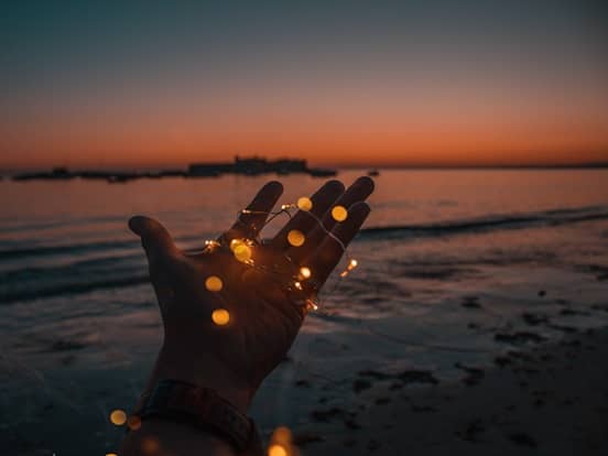 Hand hält Lichterkette über Meer mit Sonnenuntergang