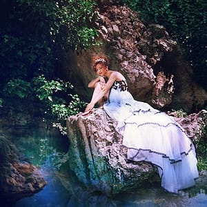 Feenhaft aussehende Frau mit Krone und in langem Kleid sitzt auf Fels am Wasser